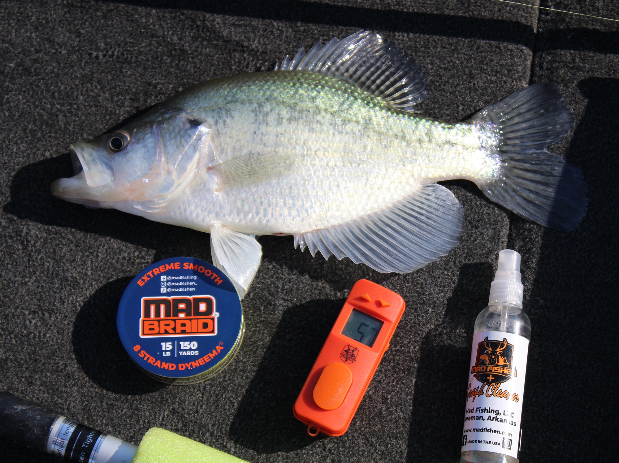 A wider view of a freshly caught crappie fish laid out on a boat deck, surrounded by essential fishing gear. Featured products include Mad Braid braided fishing line for smooth casting, The Button digital fish counter for easy catch tracking, and MadFishen Graph Cleaner for keeping electronics spotless. The ultimate setup for a successful fishing trip.