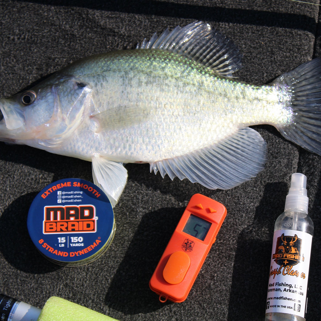 A freshly caught crappie fish displayed on a boat deck alongside top fishing essentials—Mad Braid braided fishing line, The Button digital fish counter, and MadFishen Graph Cleaner. The perfect combination for a successful fishing trip.