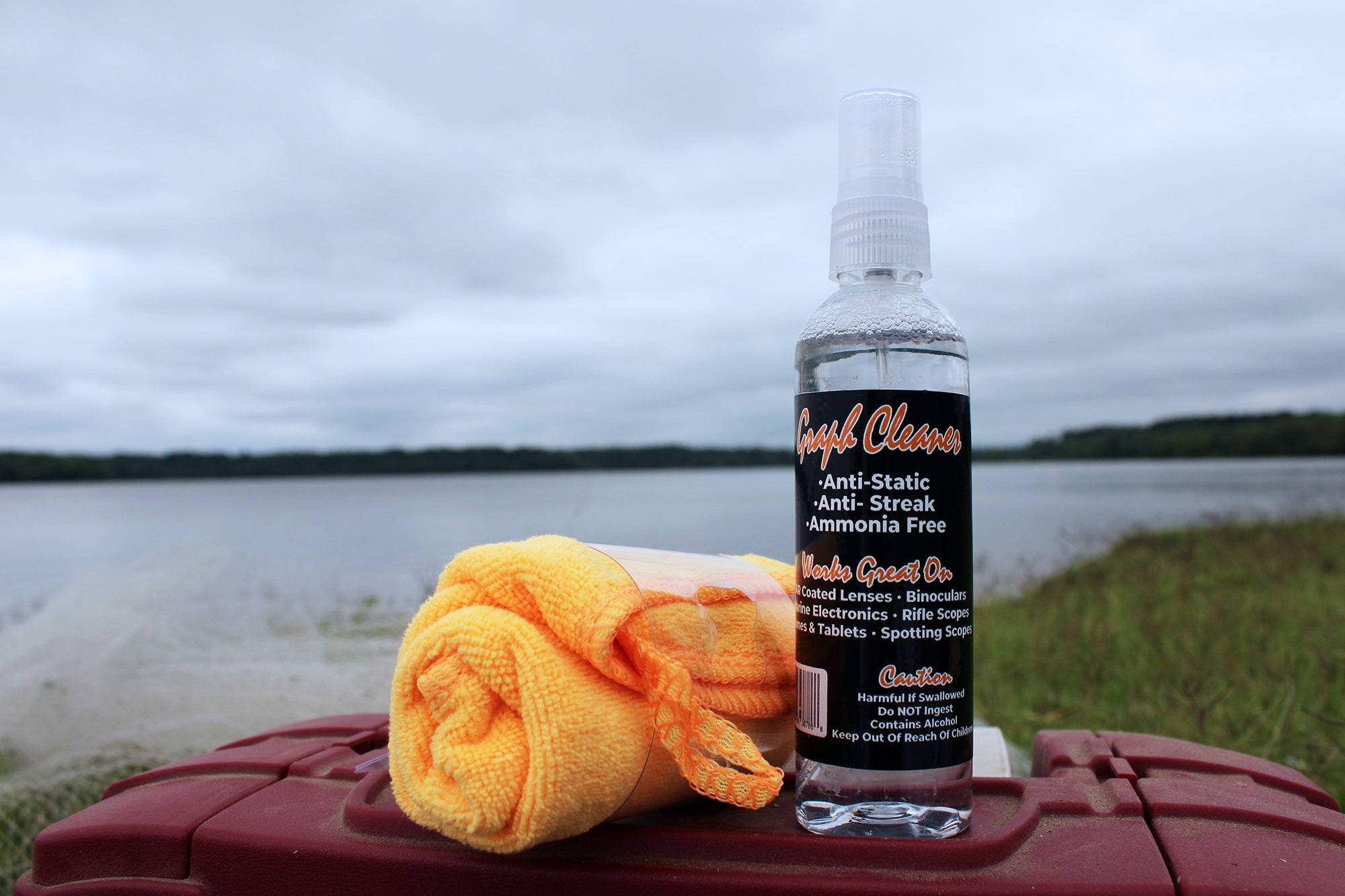 MadFishen Graph Cleaner bottle with a microfiber towel placed on a tackle box near a lake. The anti-static, streak-free screen cleaner is perfect for keeping fish finders, coated lenses, and marine electronics spotless on fishing trips.