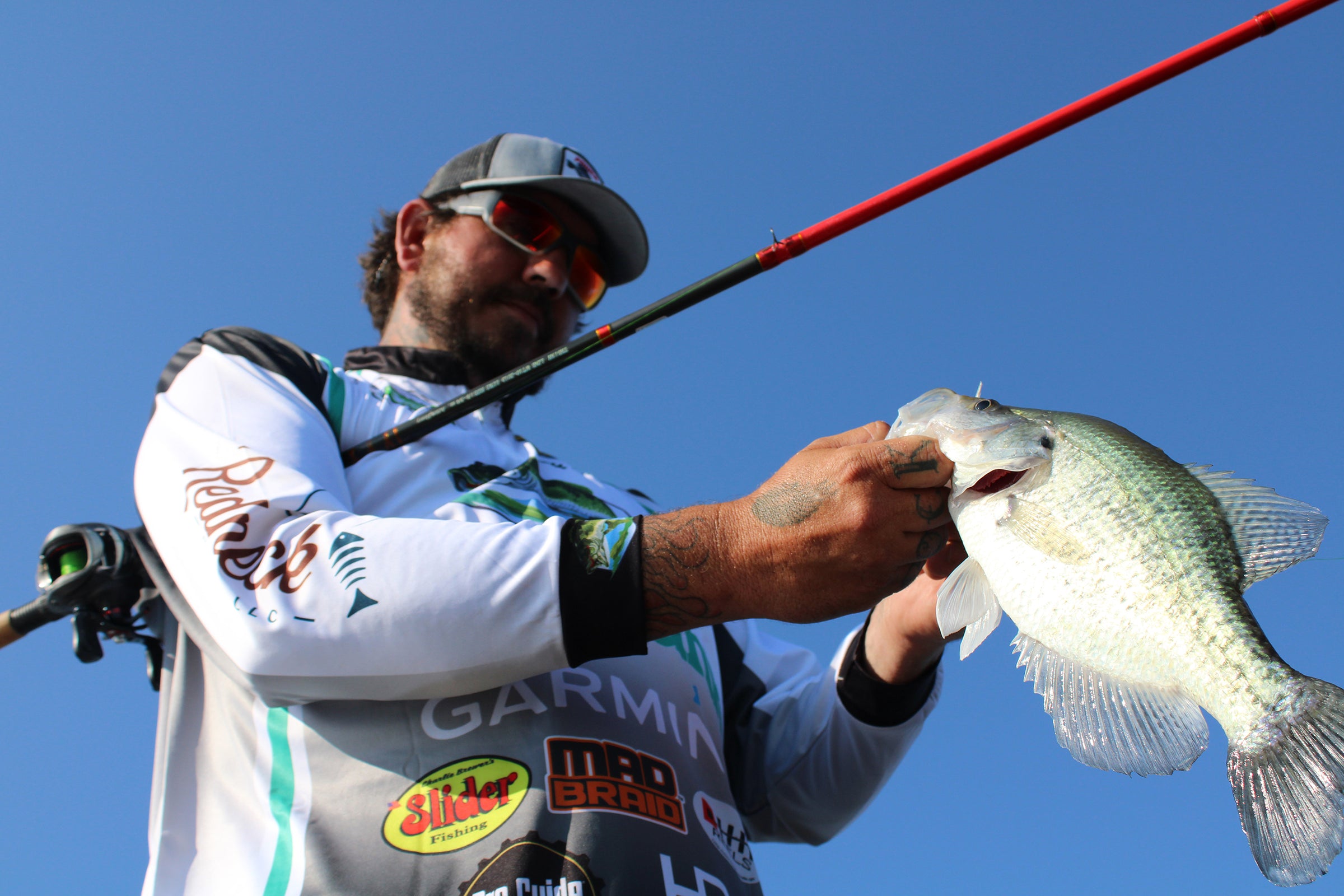 A professional angler wearing a fishing jersey holds a freshly caught crappie against a bright blue sky. He uses a rod spooled with Mad Braid braided fishing line, known for its strength and smooth casting, ensuring a reliable fishing experience.